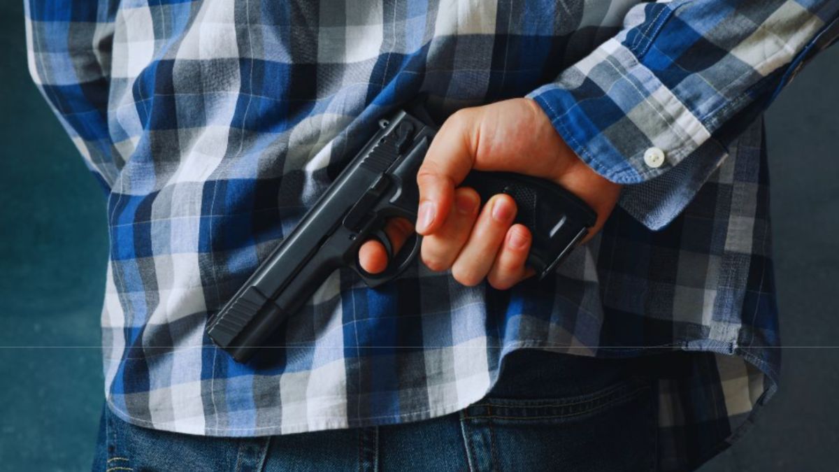 Posesión de un arma de fuego 2 Person in a blue plaid shirt holding a handgun behind their back, with the weapon pointed downward, symbolizing unlawful possession of a firearm by a convicted felon.