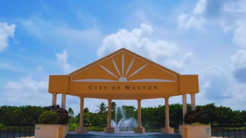 Weston, FL 2 Decorative “City of Weston” gateway sign in Weston, Florida, with a stylized sunburst design, fountain, palm trees, and a bright blue sky with scattered clouds in the background.