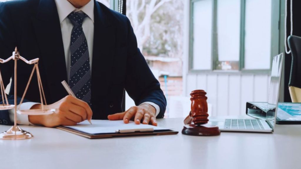 Drug crimes defense attorney in a suit writing on legal documents at a desk with scales of justice, gavel, and laptop in a Fort Lauderdale law office.