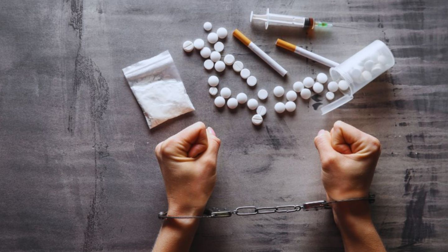 Hands in handcuffs on a gray surface in front of scattered white pills, syringes, cigarettes, and a bag of white powder, symbolizing an arrest on drug crime charges.